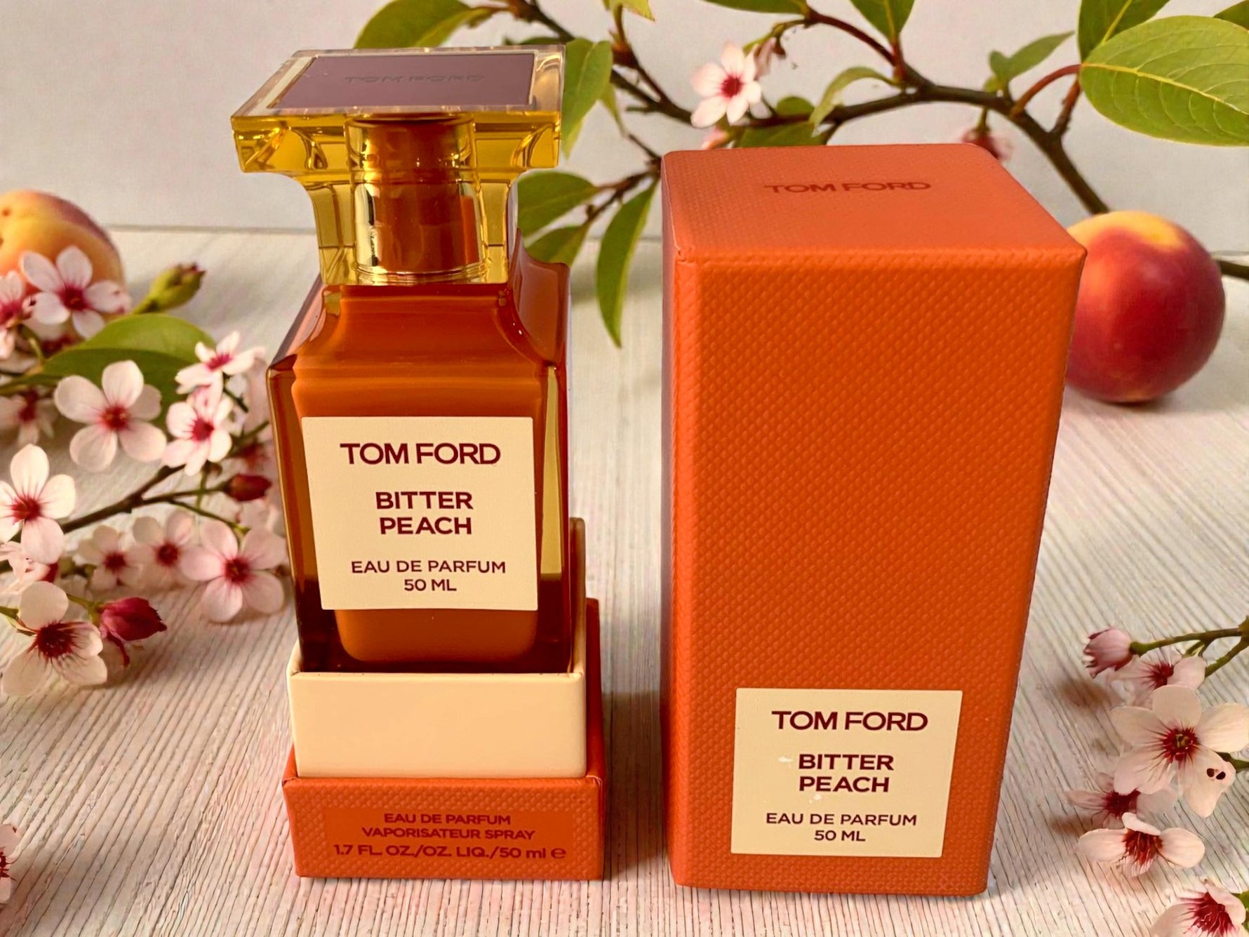 Tom Ford Bitter Peach perfume bottle and packaging on a light wooden surface with flowers and peaches